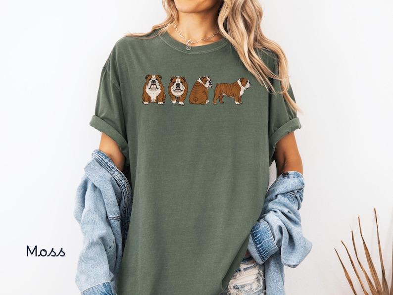 May include: A moss green t-shirt with a design of four cartoon bulldogs in different poses. The bulldogs are brown and white with black outlines. The shirt has short sleeves and a relaxed fit. The word "Moss" is printed in the bottom left corner.