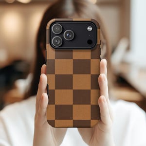 Puede incluir: Una funda de teléfono a cuadros marrones y marrón oscuro. La funda tiene un recorte para la cámara con tres lentes y un flash. La funda se sostiene frente a una persona.