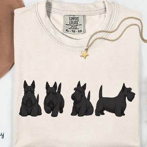 May include: Ivory-colored t-shirt featuring four black Scottish Terrier illustrations. A gold necklace with a star pendant rests on the shirt. The shirt has a Comfort Colors tag. A brown hat and flowers are in the background.