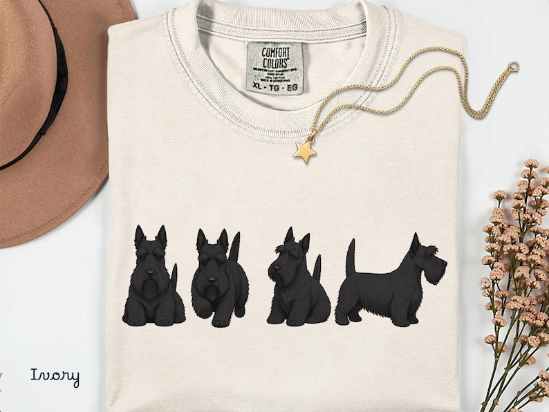 May include: Ivory-colored t-shirt featuring four black Scottish Terrier illustrations. A gold necklace with a star pendant rests on the shirt. The shirt has a Comfort Colors tag. A brown hat and flowers are in the background.