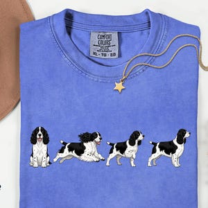 May include: A periwinkle blue t-shirt featuring a graphic print of four black and white English Springer Spaniel dogs in various poses. The shirt has a Comfort Colors brand label at the inner neckline with size XL, TG, EG printed on the tag.