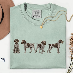 Puede incluir: Una camiseta de color verde claro con un estampado gráfico de cuatro perros de muestra alemanes de pelo corto en varias posturas. La camiseta tiene una etiqueta de la marca Comfort Colors en el cuello interior que indica la talla XL, TG, EG. La palabra Bay está impresa debajo.
