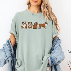 May include: A pale green t-shirt with a design of four cartoon English Bulldogs. The dogs are depicted in different poses, with brown and white markings. The shirt has a loose fit and rolled sleeves.