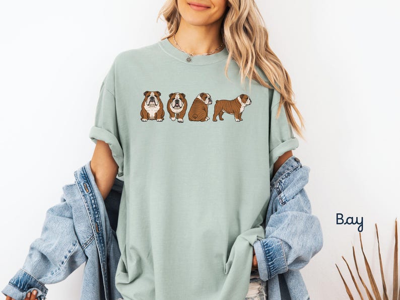 May include: A pale green t-shirt with a design of four cartoon English Bulldogs. The dogs are depicted in different poses, with brown and white markings. The shirt has a loose fit and rolled sleeves.