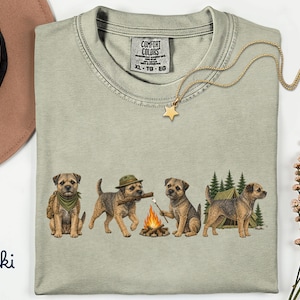 May include: A khaki t-shirt featuring a graphic of four Border Terrier dogs camping. The dogs are shown sitting, wearing a hat, holding a stick, and roasting a marshmallow over a campfire near a tent and pine trees. The shirt has a Comfort Colors label.