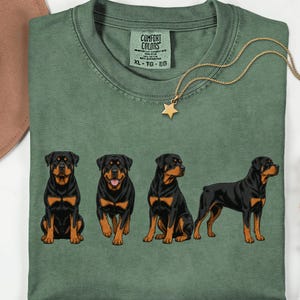Könnte beinhalten: Ein moosgrünes T-Shirt mit einem Grafikdruck von vier Rottweiler-Hunden in verschiedenen Posen. Das Shirt hat ein Comfort Colors Markenetikett am Hals, das den Text XL-TG-EG enthält. Die Hunde sind schwarz und lohfarben gezeichnet.