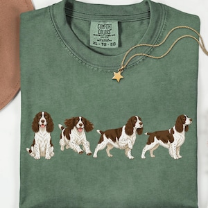 May include: A moss green crew neck t-shirt featuring a graphic print of four brown and white English Springer Spaniels in various poses. The shirt has a Comfort Colors label at the inner neckline, indicating a size XL, TG, EG.