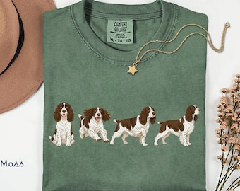 May include: A moss green crew neck t-shirt featuring a graphic print of four brown and white English Springer Spaniels in various poses. The shirt has a Comfort Colors label at the inner neckline, indicating a size XL, TG, EG.