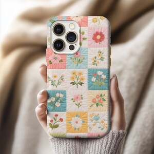 May include: A floral patterned phone case featuring a patchwork design with squares of pastel colors. Each square showcases a different flower arrangement. The case is held in a person's hand, showcasing the design.