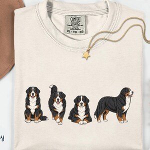 Op de afbeelding: Ivoor T-shirt met een afbeelding van vier Berner Sennenhonden in verschillende poses. Het shirt heeft een ronde hals en een label "COMFORT COLORS". Een gouden ketting met een sterhanger en een bruine hoed zijn ook zichtbaar. Maat XL-TG-EG.