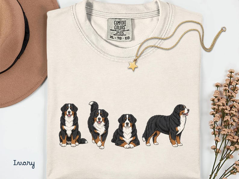 Op de afbeelding: Ivoor T-shirt met een afbeelding van vier Berner Sennenhonden in verschillende poses. Het shirt heeft een ronde hals en een label "COMFORT COLORS". Een gouden ketting met een sterhanger en een bruine hoed zijn ook zichtbaar. Maat XL-TG-EG.