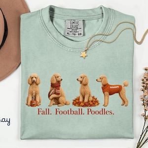 May include: A light green t-shirt featuring a graphic of four apricot poodles in autumn-themed poses, including one with a mug, one with a scarf and football, one in leaves, and one in a football jersey. The text reads Fall. Football. Poodles.