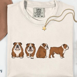 May include: Ivory t-shirt with four brown and white bulldog illustrations. The shirt has a crew neck and a tag that reads "COMFORT COLORS". A gold necklace with a star pendant is draped across the shirt. The shirt is XL.