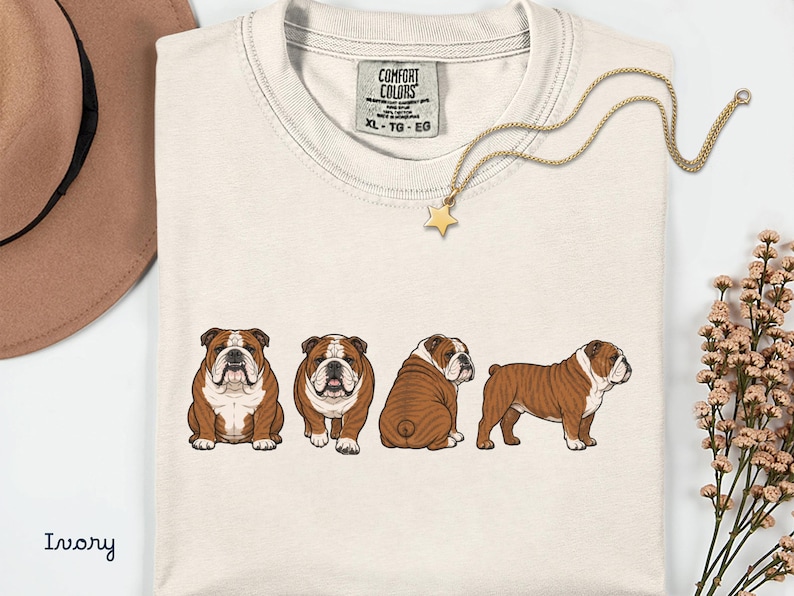 May include: Ivory t-shirt with four brown and white bulldog illustrations. The shirt has a crew neck and a tag that reads "COMFORT COLORS". A gold necklace with a star pendant is draped across the shirt. The shirt is XL.