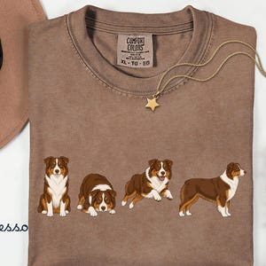 May include: An espresso-colored t-shirt featuring a graphic print of four Australian Shepherd dogs in various poses. The shirt has a crew neckline and a Comfort Colors brand label on the inside collar. A gold-toned star pendant necklace is placed on top.