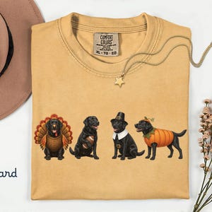 May include: A mustard yellow crew neck t-shirt featuring a graphic of four black Labrador dogs. The dogs are dressed as a turkey, a football player, a pilgrim, and a pumpkin. The shirt has a Comfort Colors brand label on the inside neck.