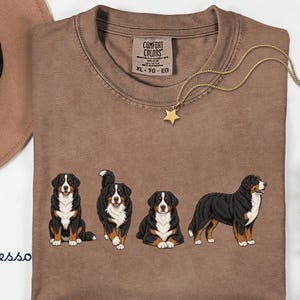 Op de afbeelding: Espresso-kleurig T-shirt met een afbeelding van vier Berner Sennenhonden in verschillende poses. Het shirt heeft een ronde hals en een label met de tekst "Comfort Colors". Een gouden sterrenhangerketting en een bruine hoed staan ook op de afbeelding.