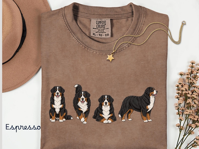 Op de afbeelding: Espresso-kleurig T-shirt met een afbeelding van vier Berner Sennenhonden in verschillende poses. Het shirt heeft een ronde hals en een label met de tekst "Comfort Colors". Een gouden sterrenhangerketting en een bruine hoed staan ook op de afbeelding.