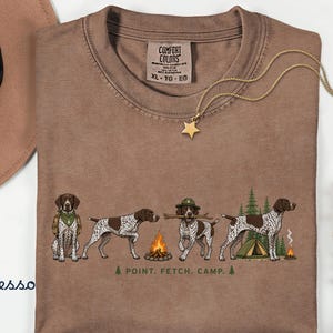 May include: A brown short-sleeved t-shirt featuring a graphic of four German Shorthaired Pointer dogs in outdoor settings. The text reads POINT. FETCH. CAMP. The shirt has a Comfort Colors label in the neck and is size XL.