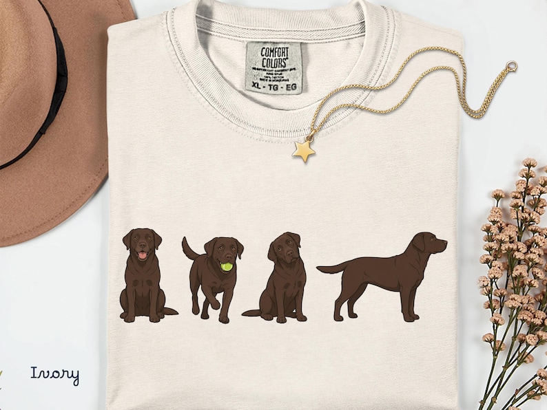 K&ouml;nnte beinhalten: Ein elfenbeinfarbenes T-Shirt mit einem Grafikdruck von vier schokoladenbraunen Labrador-Retrievern in verschiedenen Posen. Das T-Shirt hat ein Comfort Colors Markenetikett am Halsausschnitt, das die Gr&ouml;&szlig;e XL-TG-EG und 100% Baumwolle angibt.