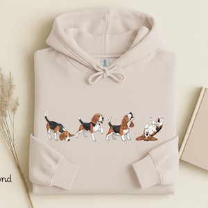 Könnte beinhalten: Ein sandfarbener Kapuzenpullover mit einem Grafikdruck von vier Beagle-Hunden in verschiedenen Posen. Der Hoodie verfügt über eine Kapuze mit Kordelzug sowie lange Ärmel mit gerippten Bündchen. Die Hunde sind in Braun, Schwarz und Weiß.