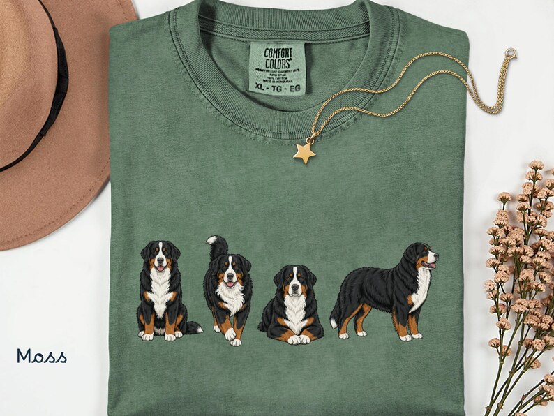 Op de afbeelding: Mosgroen t-shirt met vier illustraties van Berner Sennenhonden. Het shirt heeft een ronde hals en korte mouwen. Een goudkleurige ketting met een sterhanger ligt op het shirt. Het shirt is gelabeld "Comfort Colors" met maat XL.
