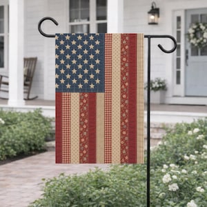 May include: A patriotic garden flag featuring the American flag design. The flag has a blue square with beige stars, red and beige stripes, and various patterns. The flag is hanging on a black metal stand in a garden setting.