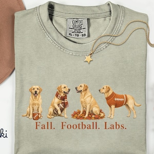 May include: A khaki crew neck t-shirt featuring a graphic of four yellow Labrador retrievers in autumn-themed attire and accessories. Below the dogs, the text reads "Fall. Football. Labs." The shirt has a Comfort Colors brand label at the neck.