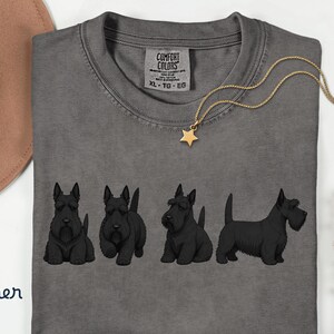 May include: A gray Comfort Colors t-shirt featuring a row of black Scottish Terrier dog illustrations. A gold necklace with a star pendant rests on the shirt. The shirt has a tag that reads "XL - TG - EG".