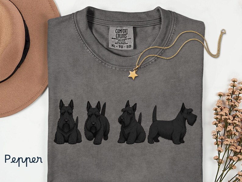 May include: A gray Comfort Colors t-shirt featuring a row of black Scottish Terrier dog illustrations. A gold necklace with a star pendant rests on the shirt. The shirt has a tag that reads "XL - TG - EG".
