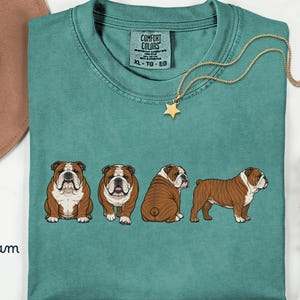 May include: Seafoam green t-shirt with four English bulldog illustrations in different poses. The shirt has a crew neck and a tag that reads "Comfort Colors". A gold star pendant hangs from a delicate chain. The word "Seafoam" is written in cursive.