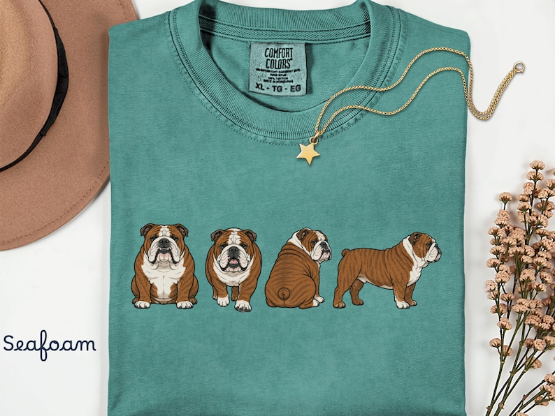 May include: Seafoam green t-shirt with four English bulldog illustrations in different poses. The shirt has a crew neck and a tag that reads "Comfort Colors". A gold star pendant hangs from a delicate chain. The word "Seafoam" is written in cursive.