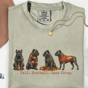 Może przedstawiać: Koszulka z okrągłym dekoltem w kolorze khaki z grafiką przedstawiającą cztery psy rasy Cane Corso. Psy są ukazane z jesiennymi akcesoriami, takimi jak kubek, szalik i sweter. Poniższy tekst brzmi: Fall. Football. Cane Corso.