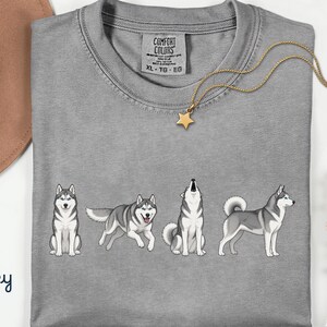 May include: A grey Comfort Colors t-shirt featuring a graphic of four cartoon huskies in different poses. A gold necklace with a star pendant is draped across the shirt. The word "Grey" is written in cursive in the bottom left corner.