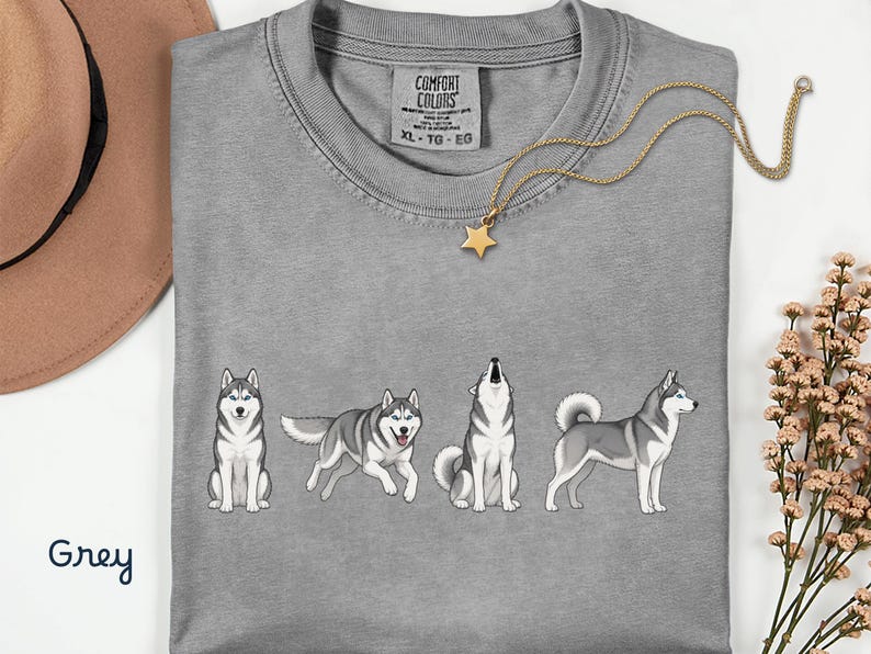 May include: A grey Comfort Colors t-shirt featuring a graphic of four cartoon huskies in different poses. A gold necklace with a star pendant is draped across the shirt. The word "Grey" is written in cursive in the bottom left corner.