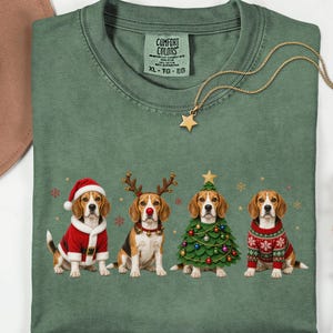 Könnte beinhalten: Ein moosgrünes T-Shirt mit Rundhalsausschnitt und einem Aufdruck von vier Beagle-Hunden in festlicher Kleidung, darunter ein Weihnachtsmannkostüm, ein Rentiergeweih mit roter Nase, ein Weihnachtsbaumkostüm und ein gemusterter Weihnachtspullover.