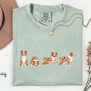 May include: A light green crew neck t-shirt featuring a graphic print of four Australian Shepherd dogs in various poses. The shirt has a Comfort Colors brand label at the inner neckline with size XL-TG-EG. The word Bay is printed in blue.