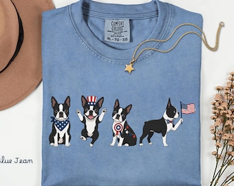 May include: Blue jean colored t-shirt featuring four cartoon Boston Terriers in patriotic attire. One dog wears a bandana, another a hat, a third a shield, and the last holds an American flag. A gold star necklace is draped on the shirt.