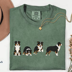 May include: A moss green crew neck t-shirt featuring a graphic print of four Australian Shepherd dogs in various poses. The shirt has a Comfort Colors brand label at the inner neckline, indicating size XL. The fabric has a soft, washed appearance.