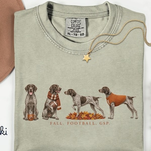 Może przedstawiać: Koszulka w kolorze khaki z metką Comfort Colors w rozmiarze XL. Na koszulce znajduje się grafika przedstawiająca cztery wyżły niemieckie krótkowłose w różnych pozach z piłkami do futbolu i jesiennymi liśćmi oraz nadrukowany poniżej napis FALL. FOOTBALL. GSP.