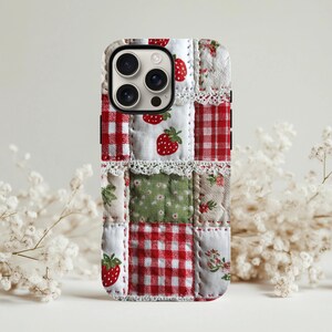 Puede incluir: Funda de teléfono con patchwork con cuadrados de tela de cuadros rojos, estampados florales y de fresas. La funda tiene un ribete de encaje blanco y un borde negro, creando una estética artesanal. Diseñada para un smartphone.