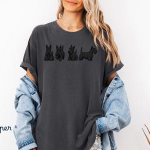 May include: Dark gray t-shirt featuring a line of four black Scottish Terrier dog illustrations. The shirt has a relaxed fit and rolled sleeves. The word "Pepper" is written in a script font on the bottom left.