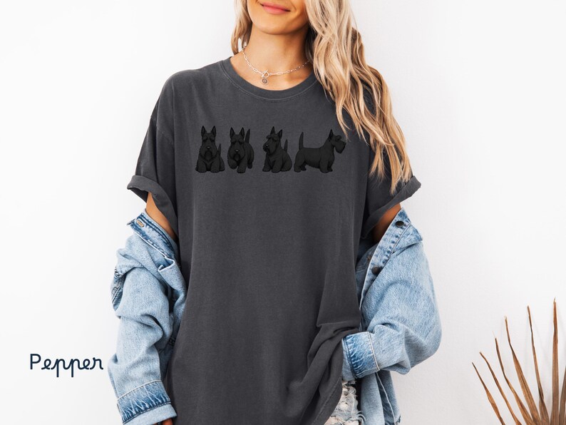 May include: Dark gray t-shirt featuring a line of four black Scottish Terrier dog illustrations. The shirt has a relaxed fit and rolled sleeves. The word "Pepper" is written in a script font on the bottom left.