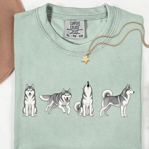 May include: A light green t-shirt featuring four cartoon huskies in different poses. The shirt has a Comfort Colors tag and a gold star pendant necklace. The word "Bay" is written in cursive on the bottom left.