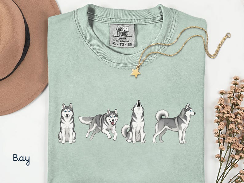 May include: A light green t-shirt featuring four cartoon huskies in different poses. The shirt has a Comfort Colors tag and a gold star pendant necklace. The word "Bay" is written in cursive on the bottom left.