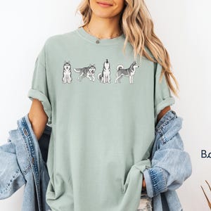 May include: A light green t-shirt featuring a line of cartoon huskies in various poses. The shirt is a solid color and has a relaxed fit. The word "Bay" is written in a script font in the lower right corner.