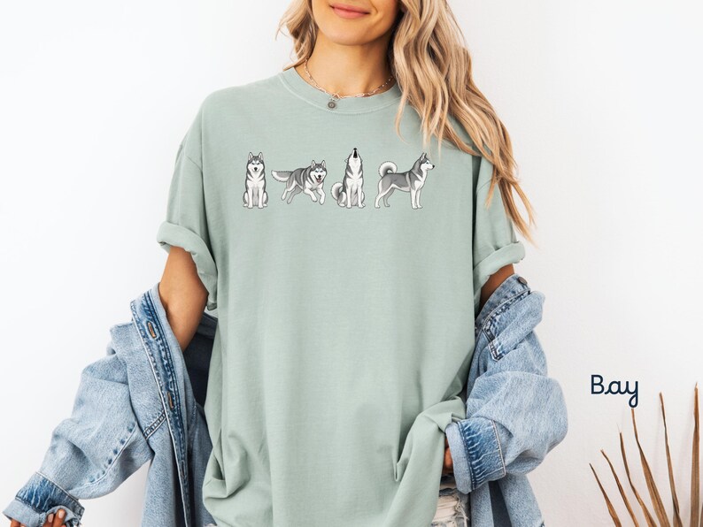 May include: A light green t-shirt featuring a line of cartoon huskies in various poses. The shirt is a solid color and has a relaxed fit. The word "Bay" is written in a script font in the lower right corner.