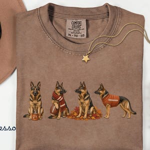 May include: An espresso-colored t-shirt featuring a graphic of four German Shepherd dogs. One dog sits with a mug, one holds a football while wearing a scarf, one sits in autumn leaves, and one wears a football-themed sweater. The shirt has a Comfort Colors label.