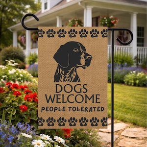 Może przedstawiać: Brązowa flaga ogrodowa z juty z czarną sylwetką psa i obramowaniem z odcisków łap. Na fladze widnieje napis "DOGS WELCOME PEOPLE TOLERATED" w czarnym tekście. Flaga jest eksponowana na czarnym metalowym paliku ogrodowym.
