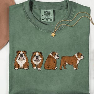 May include: A moss green t-shirt with a series of four cartoon bulldogs in different poses. The shirt has a crew neck and short sleeves. A gold-coloured necklace with a star pendant hangs on the shirt. The text "COMFORT COLORS" is visible on the tag.